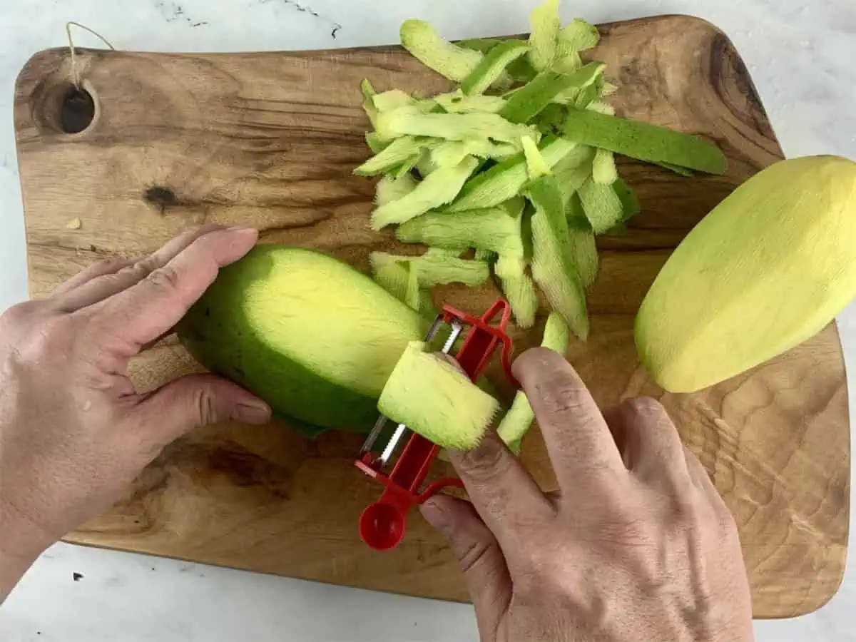 Hands peeling green mangos on a wooden board with a red y-peeler.