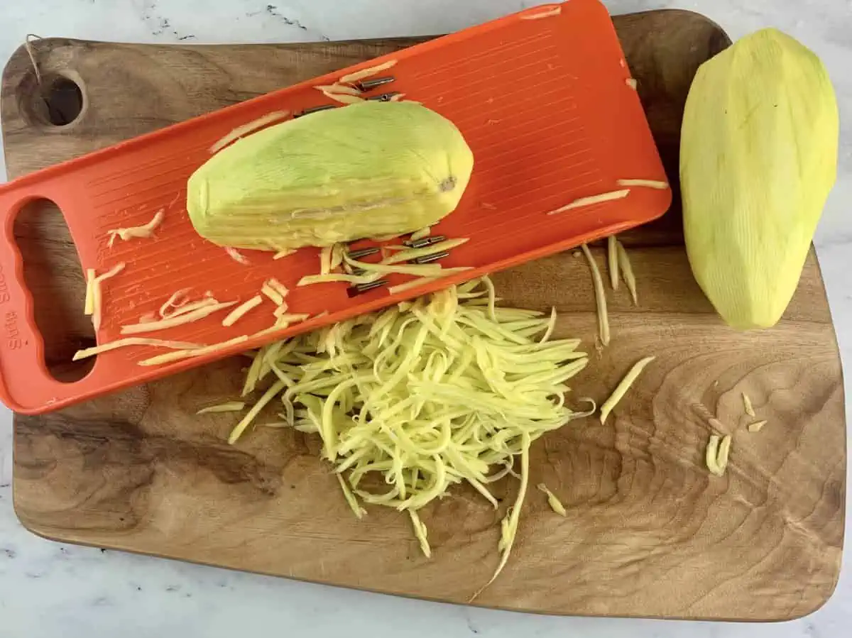 Using a mandoline to cut a green mango into julienne strips on a wooden board.