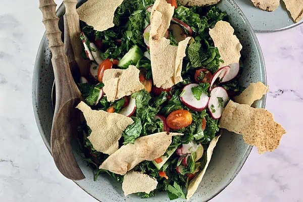 Kale Fatoush salad garnished with pita crisps in a ceramic bowl with wooden servers and more pita crisps on the side.