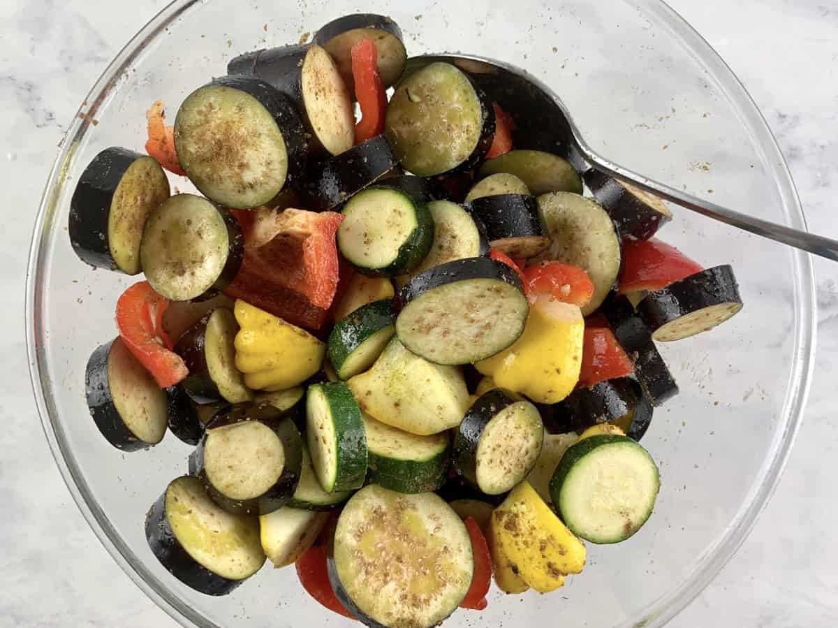 A spoon mixing summer veggies in a glass bowl with oil and spices.