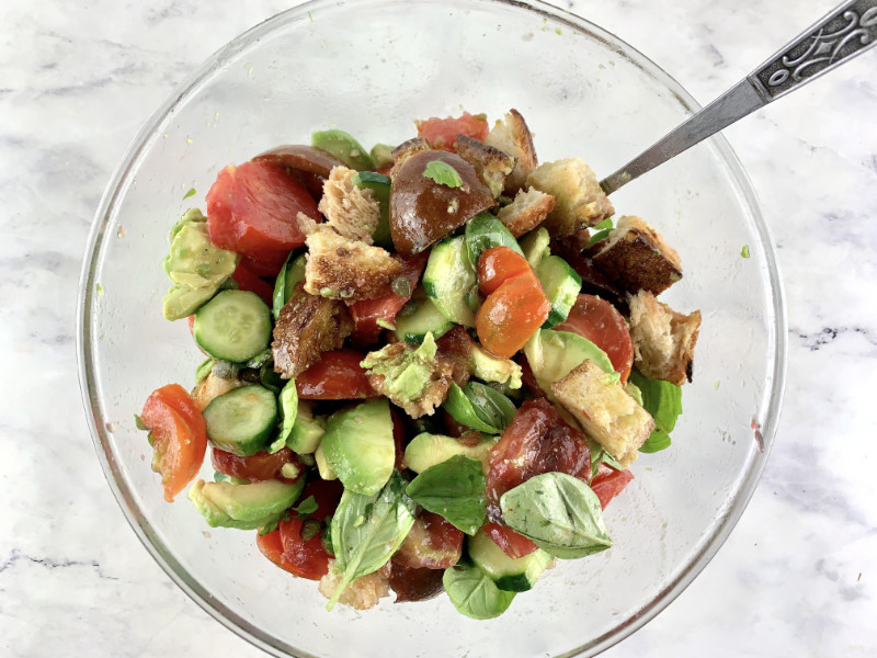 Mixing avocado Panzanella in a glass bowl with a spoon.
