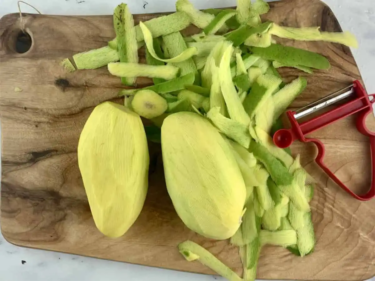 Peeled green mangos on a wooden board with a red peeler on the side.