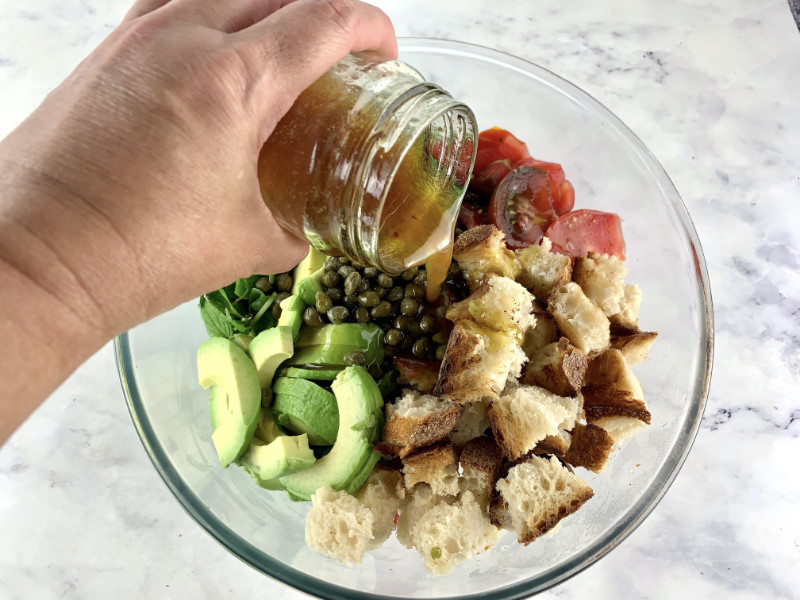 Hands pouring dressing over avocado panzanella in a glass bowl.