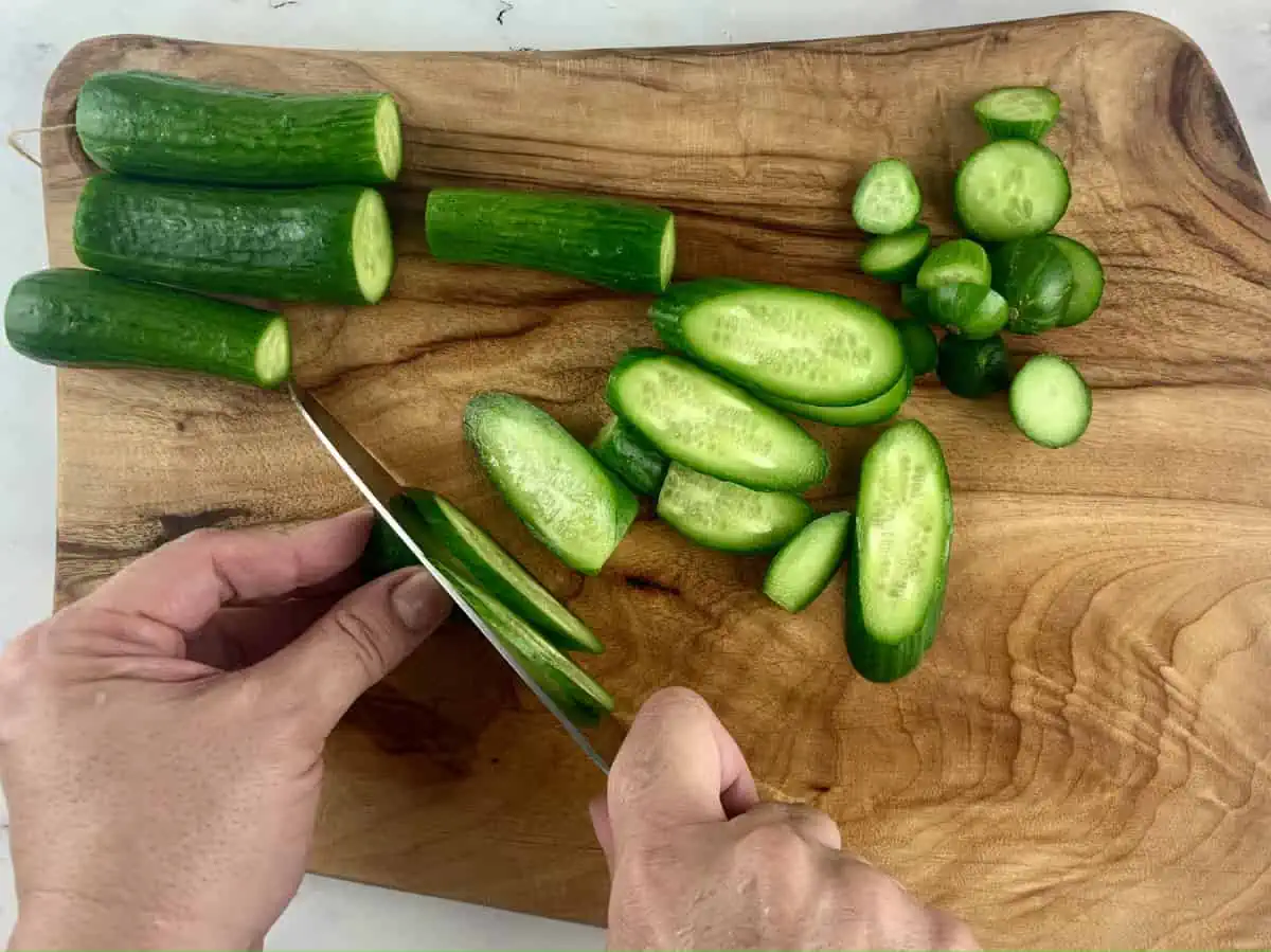 Hands slicing baby cucumbers on a wooden board with a knife.