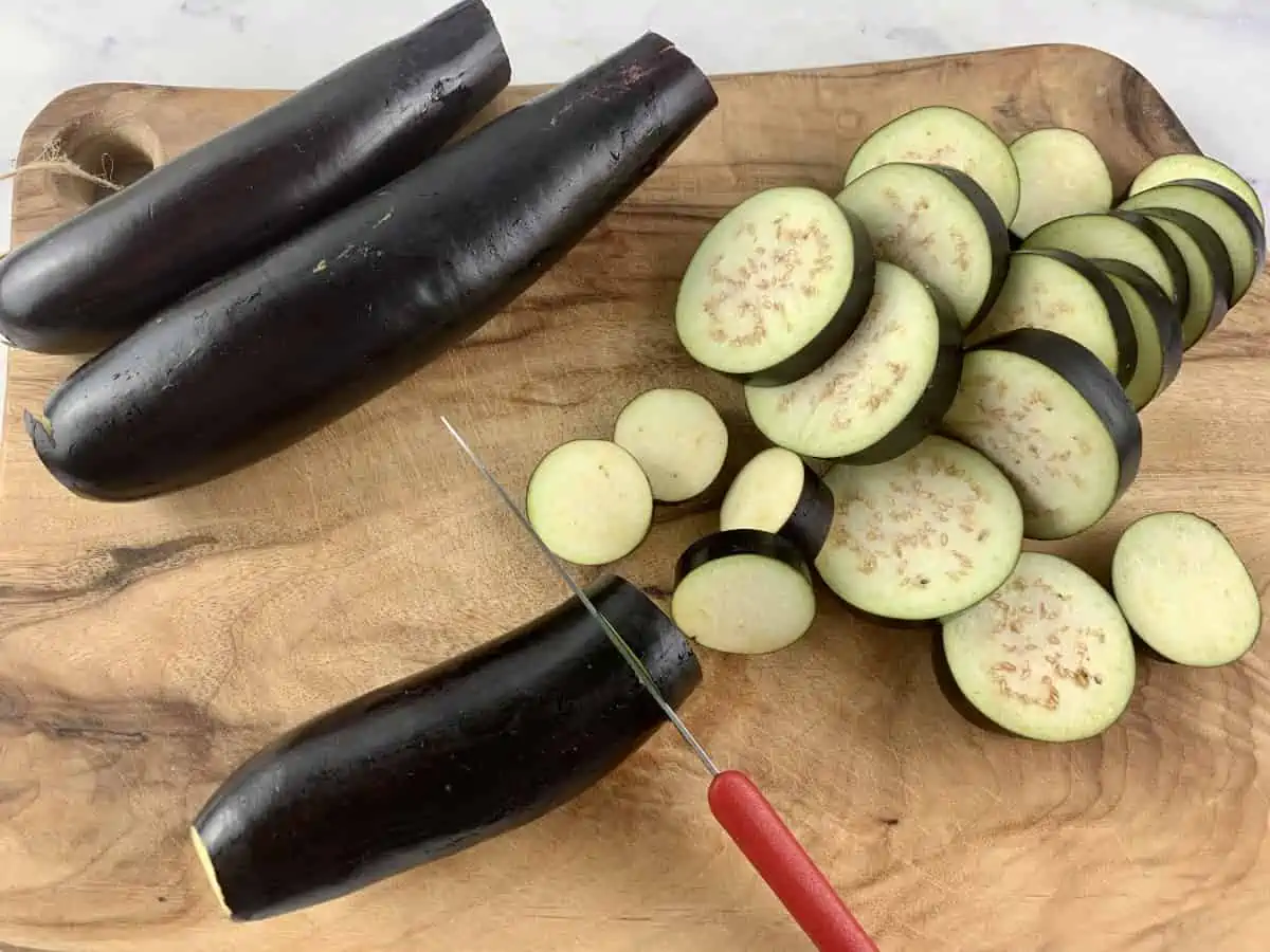 A knife slicing eggplant into rounds on a wooden board.
