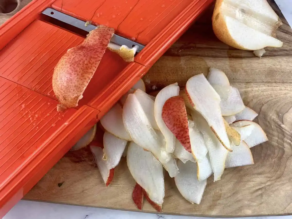 Red pears being sliced on mandoline over a wooden chopping board.