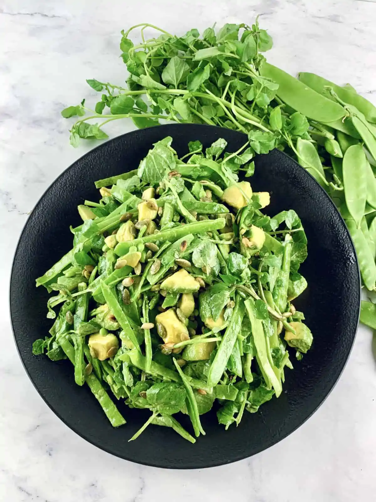 Snow pea salad on a black plate with peas and watercress on the side.