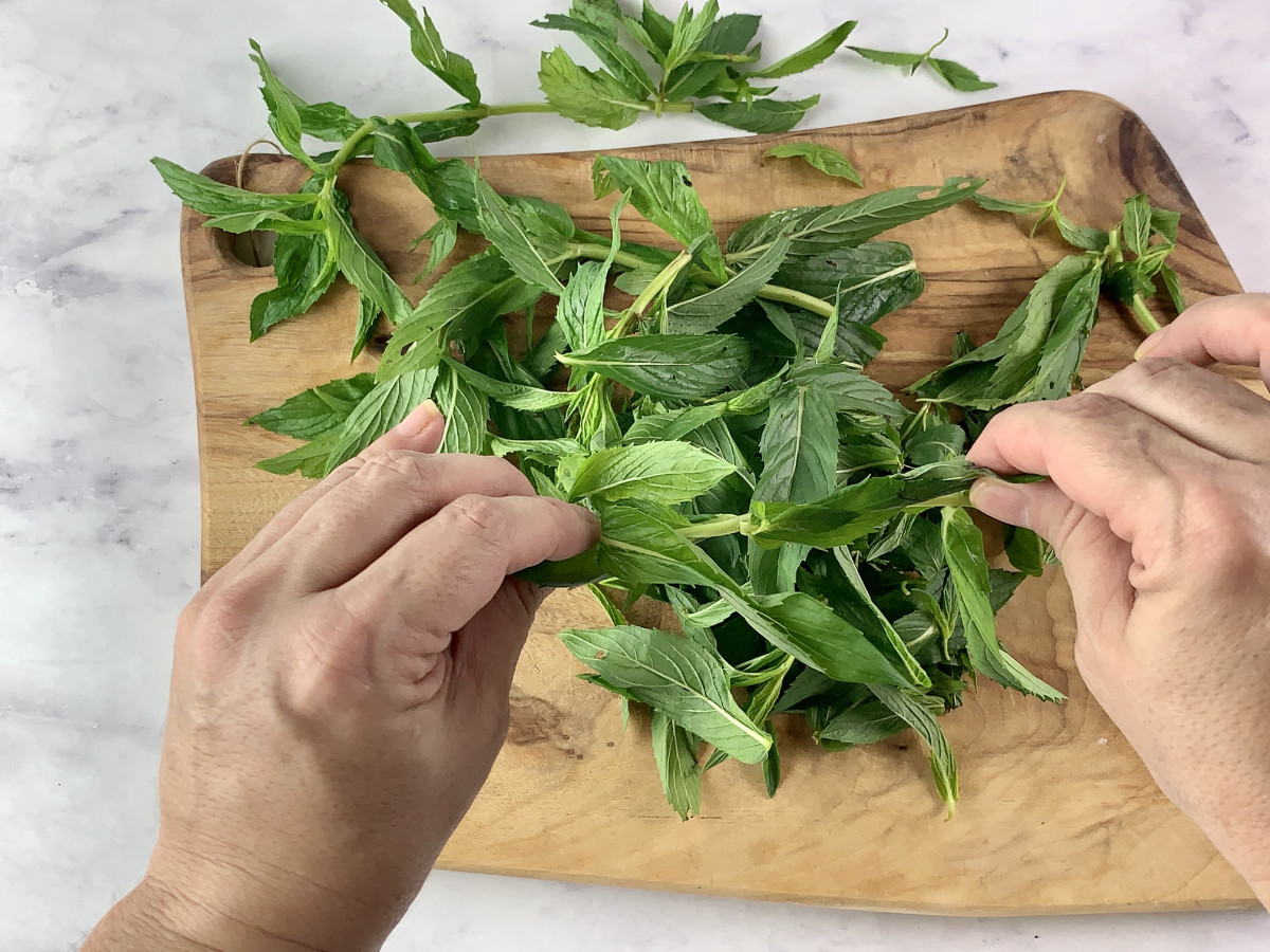 Hands stripping mint leaves from stems on a wooden board.