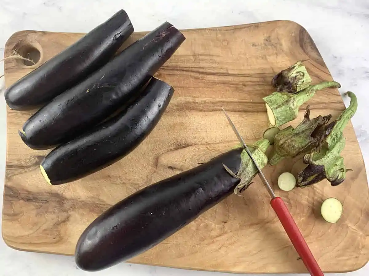 A kife trimming long eggplants on a wooden chopping board.