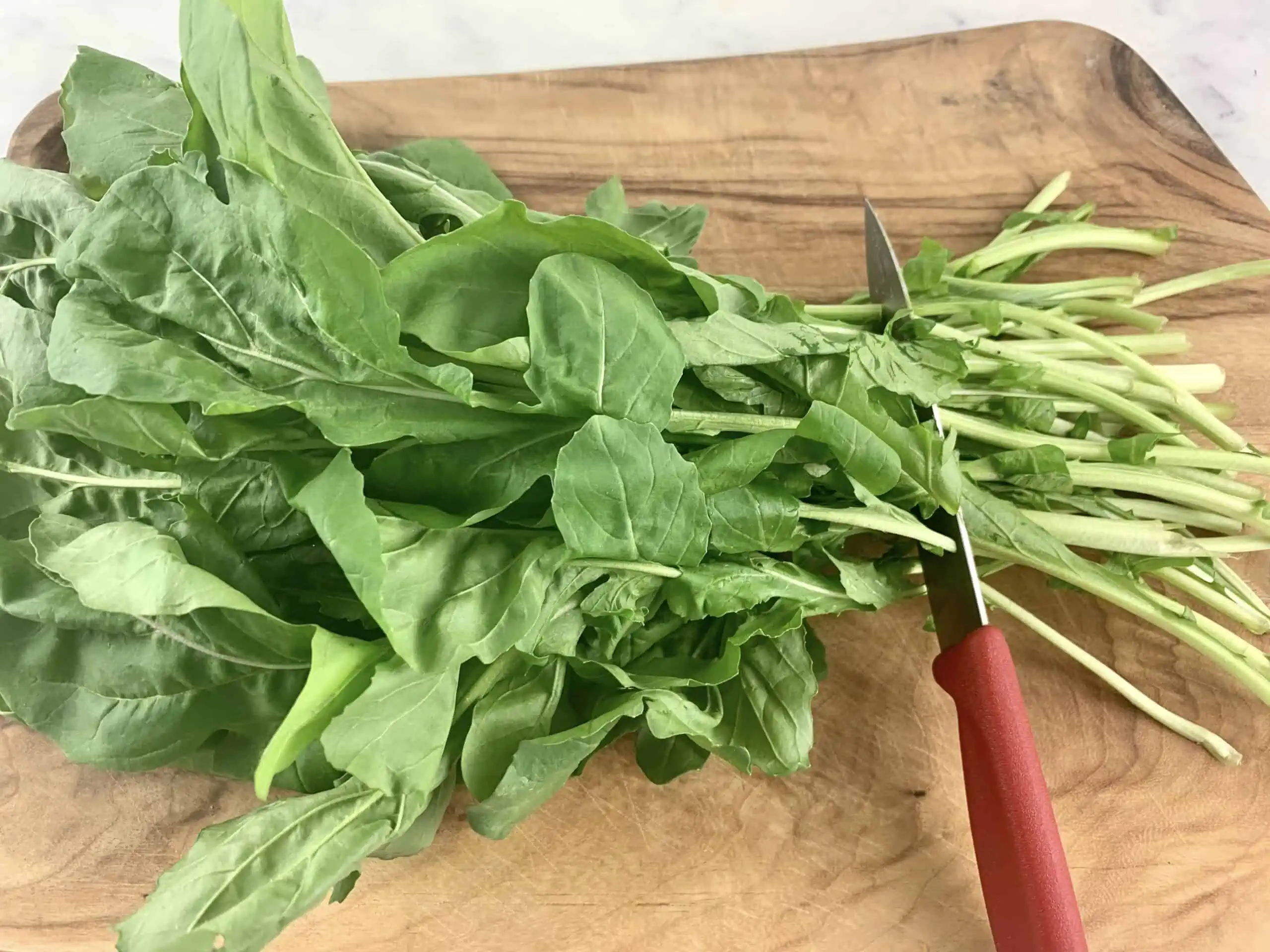 A knife trimming a bunch of rocket on.a wooden board.