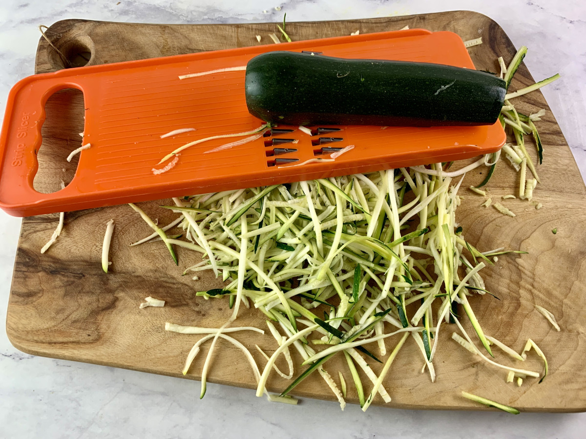 Cutting long dark green zucchini into julienne strips with v-slicer on a wooden board.