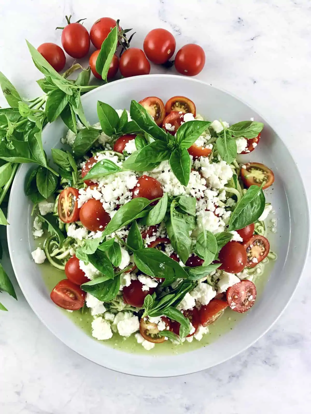 A close-up of zucchini tomato salad with feta on a white plate with basil sprigs and tomatoes on the side.