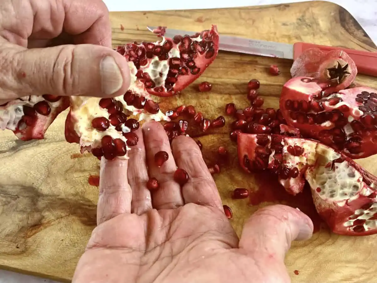 Pomegranate arils falling onto hand over a wooden board.