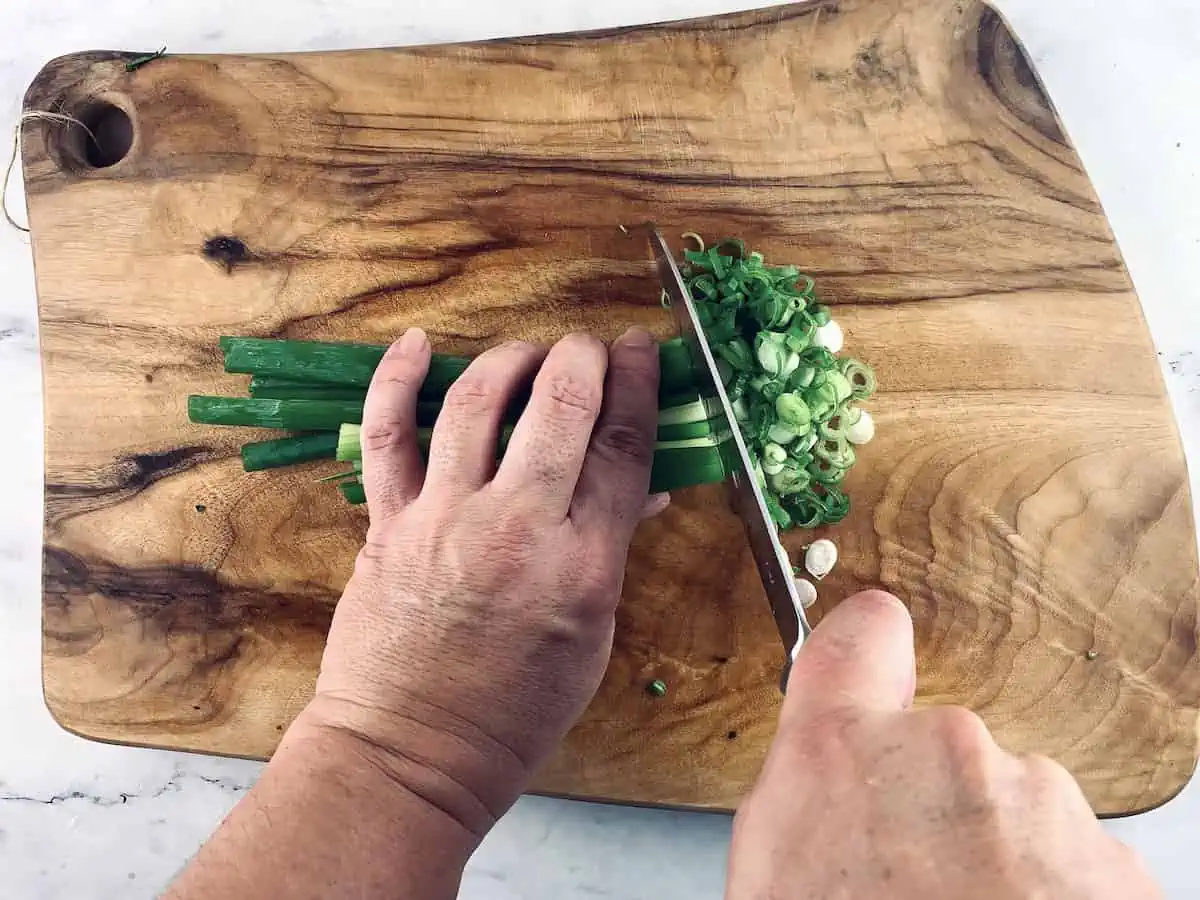 Hands chopping green onions on a wooden board with a knife.