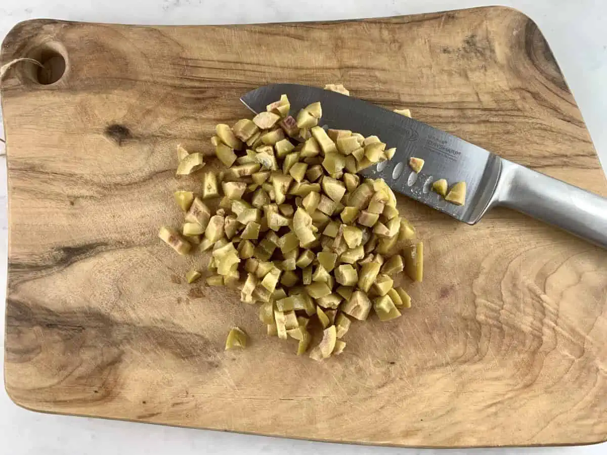 Diced green olives on a wooden board with a knife on the side.