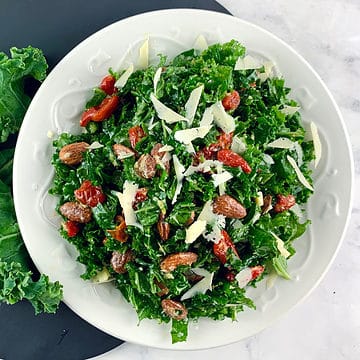 Kale lemon parmesan salad in white bowl on dark grey round board & kale leaves on the side.