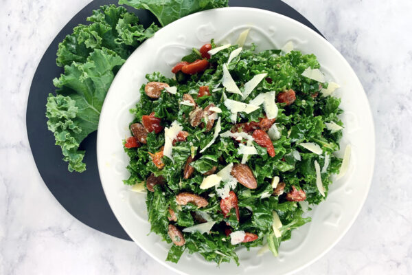 Kale lemon parmesan salad in white bowl on dark grey round board & kale leaves on the side.