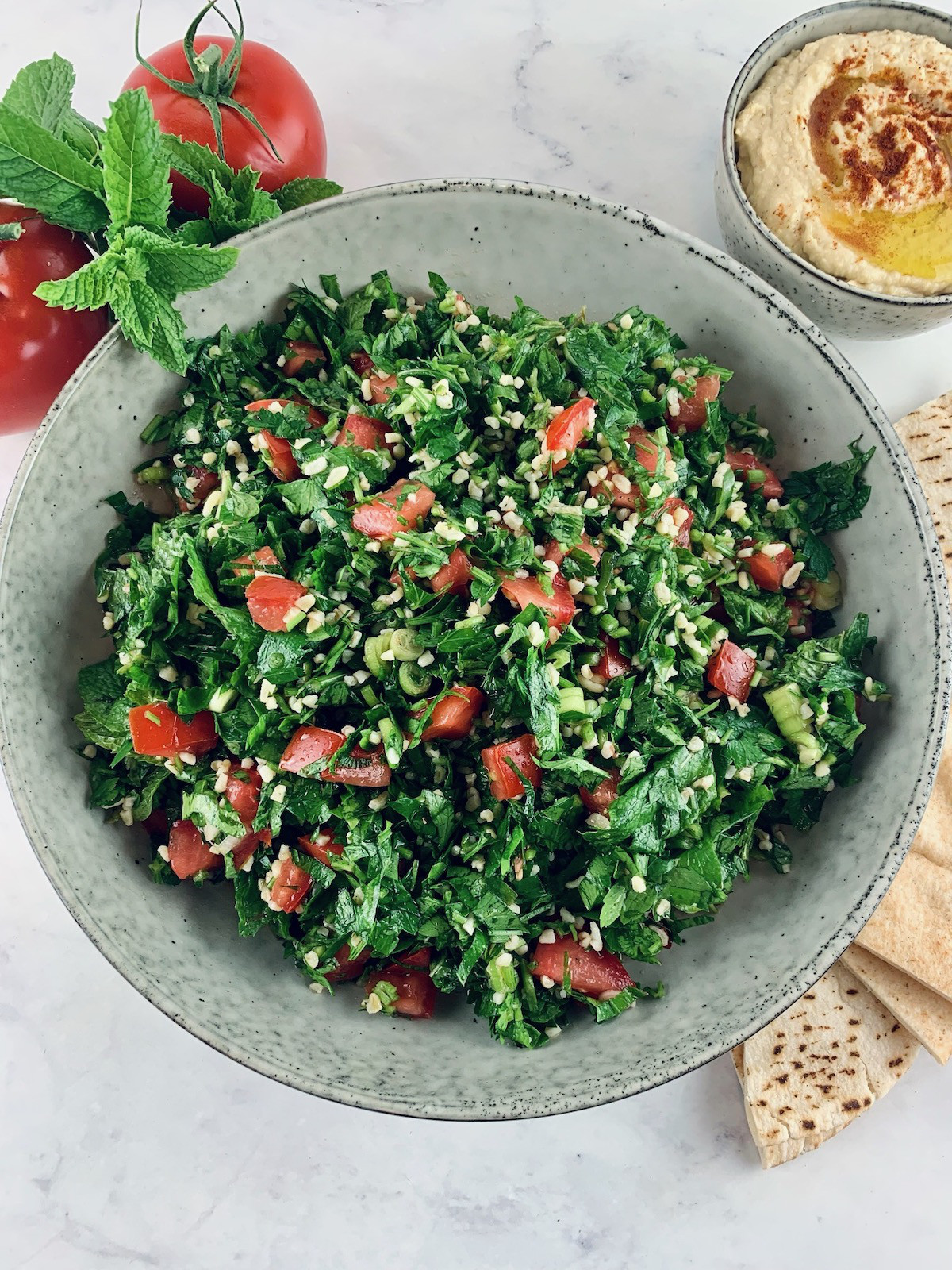 Lebanese tabbouleh in a ceramic bowl with tomatoes, hummus and pita bread on the side.