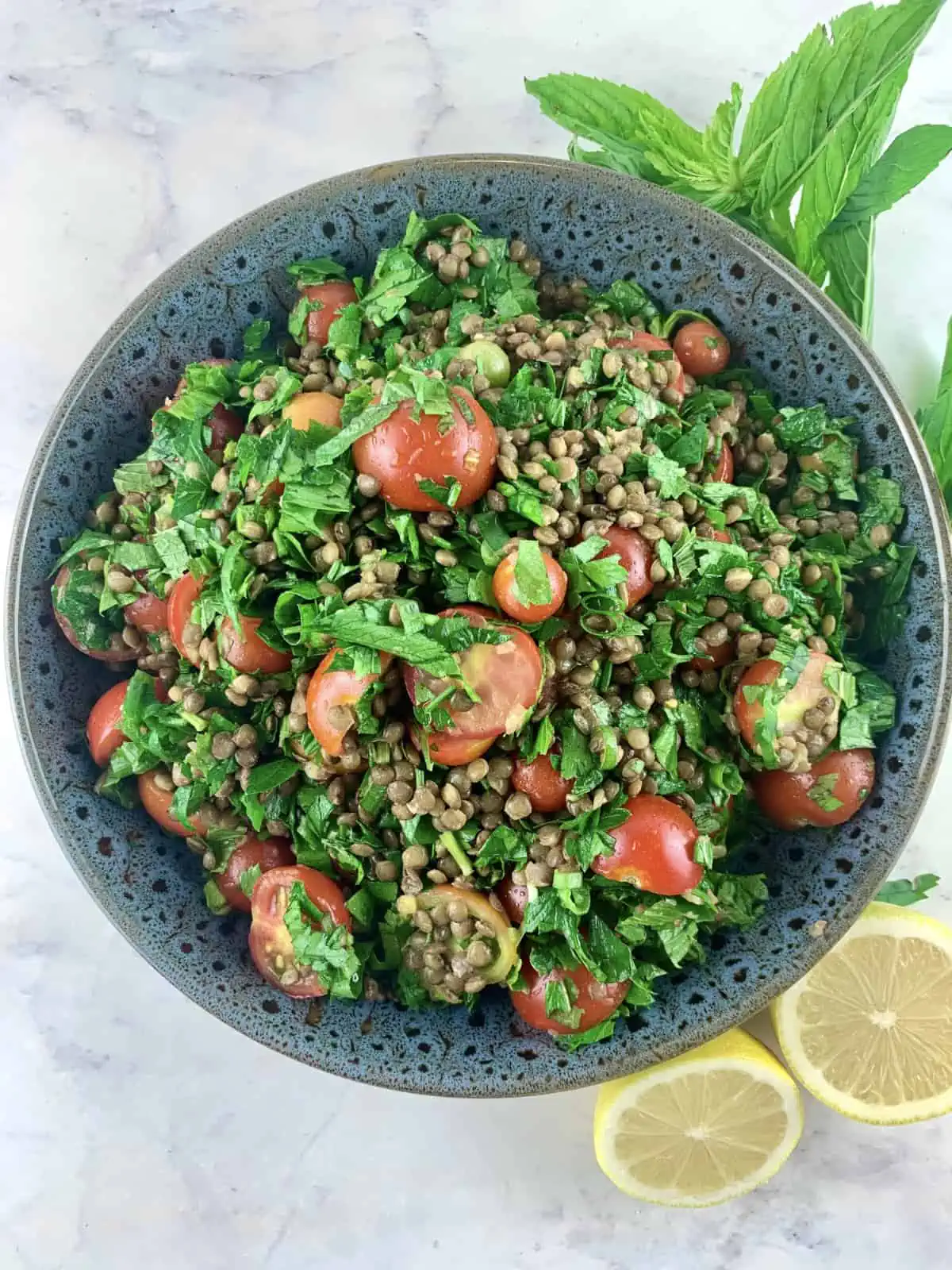 Lentil tabbouleh in a dark grey bowl with lemon halves and sprigs of mint on the side.