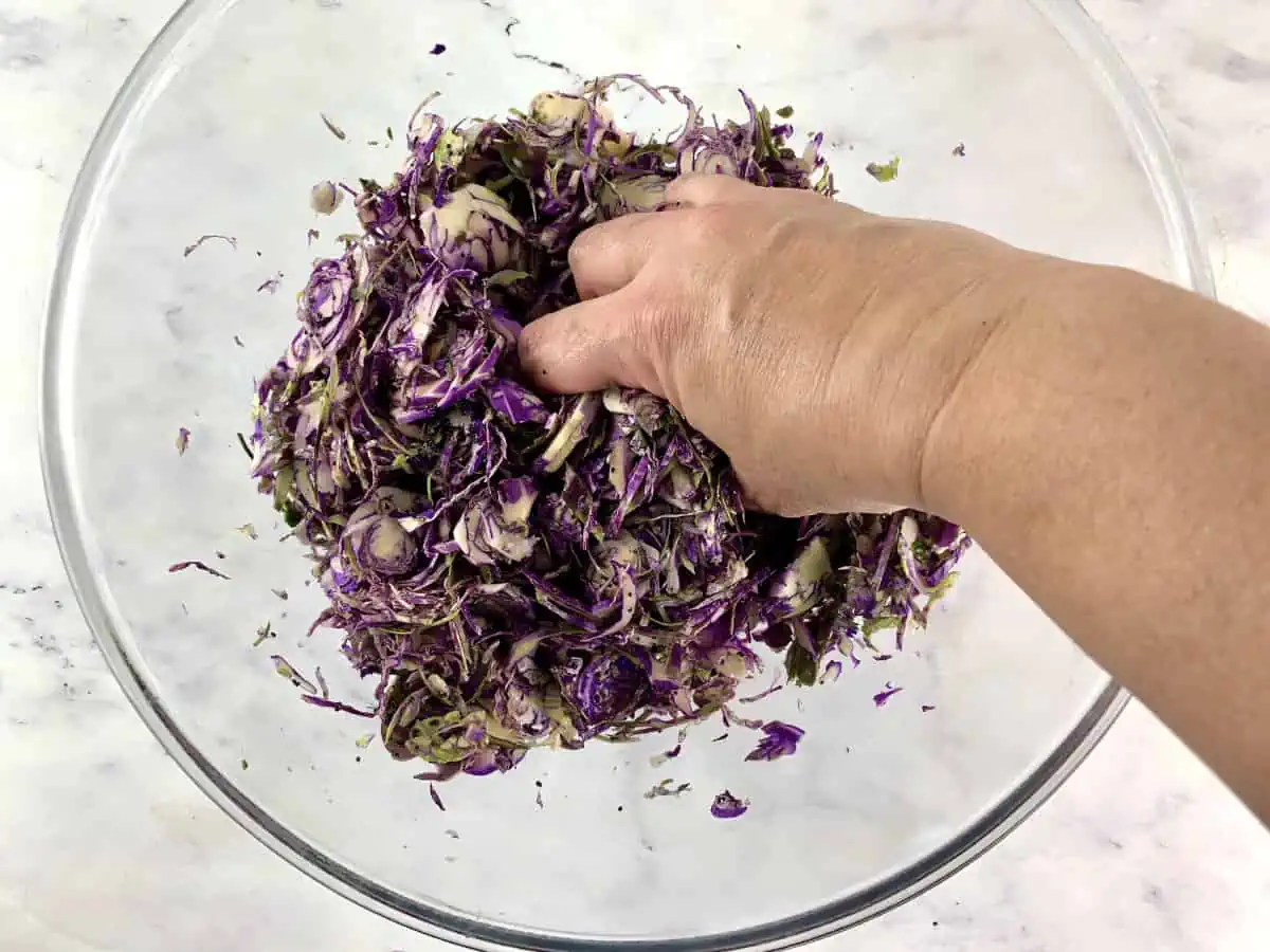 Hand massaging shredded purple brussel sprouts in a glass bowl.