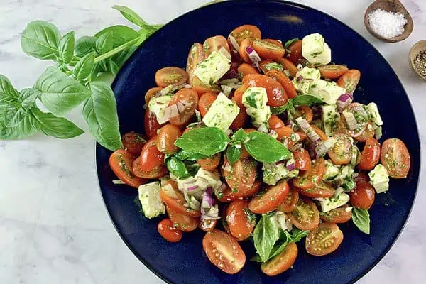 Mediterranean tomato feta salad in a dark blue bowl with basil sprig and salt and pepper on the side.