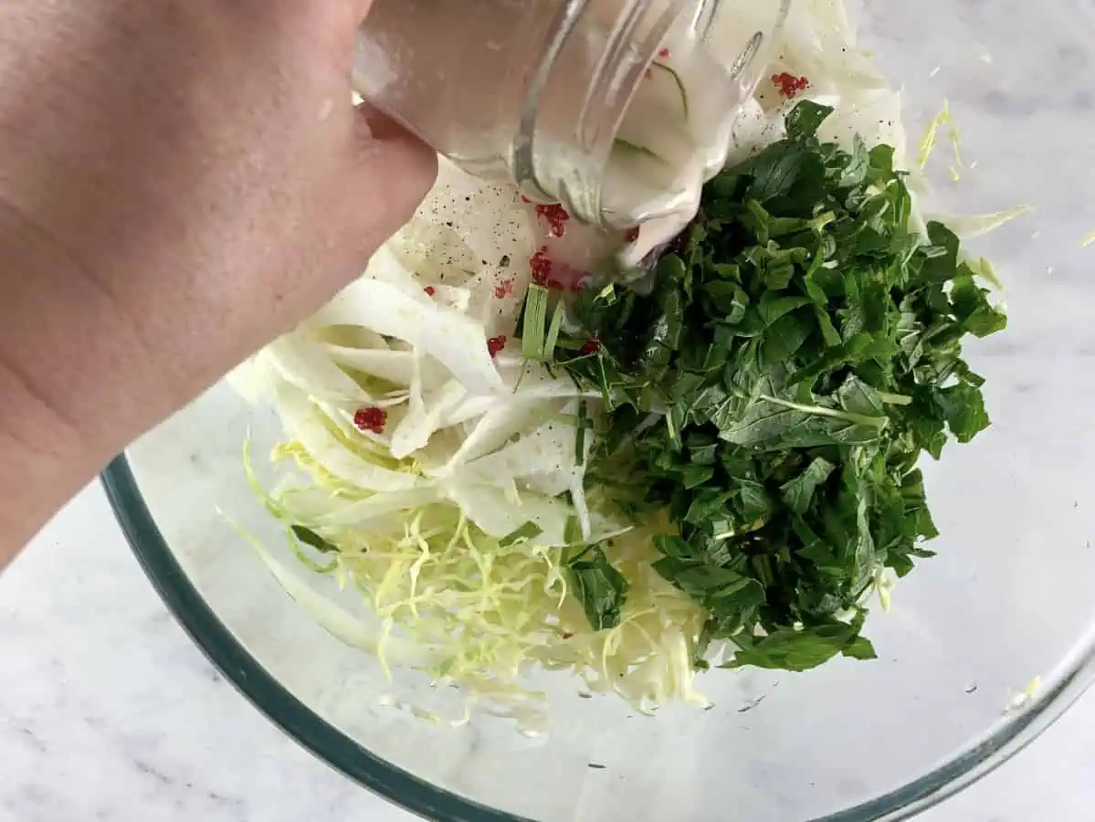 Hands pouring dressing over prepared cabbage fennel slaw ingredients.