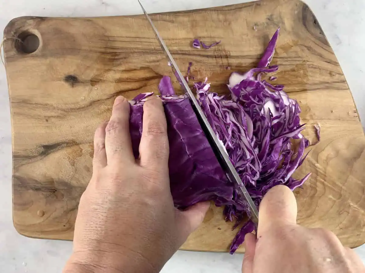 Hands shredding red cabbage on a wooden board.