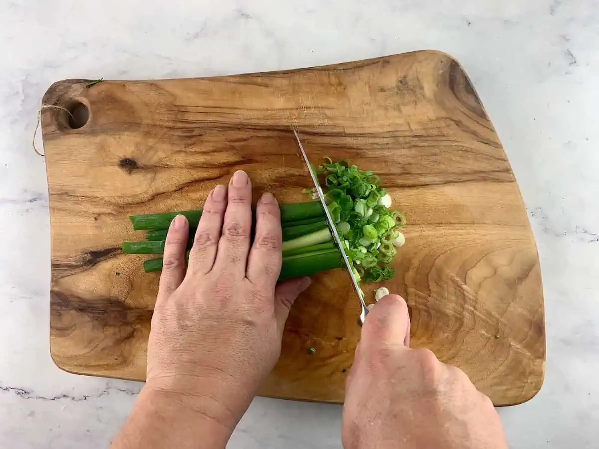 Hands slicing green onions on a wooden board.