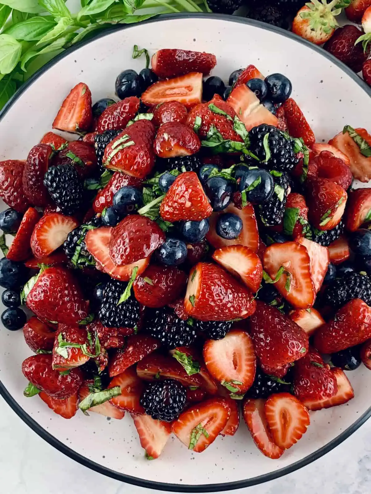 A close up of Summer berry salad on a white plate with basil sprigs and berries on the side.