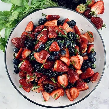 Summer berry salad on a white plate with basil sprigs and berries on the side.