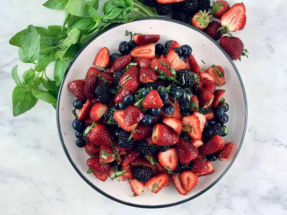 Summer berry salad on a white plate with basil sprigs and berries on the side.