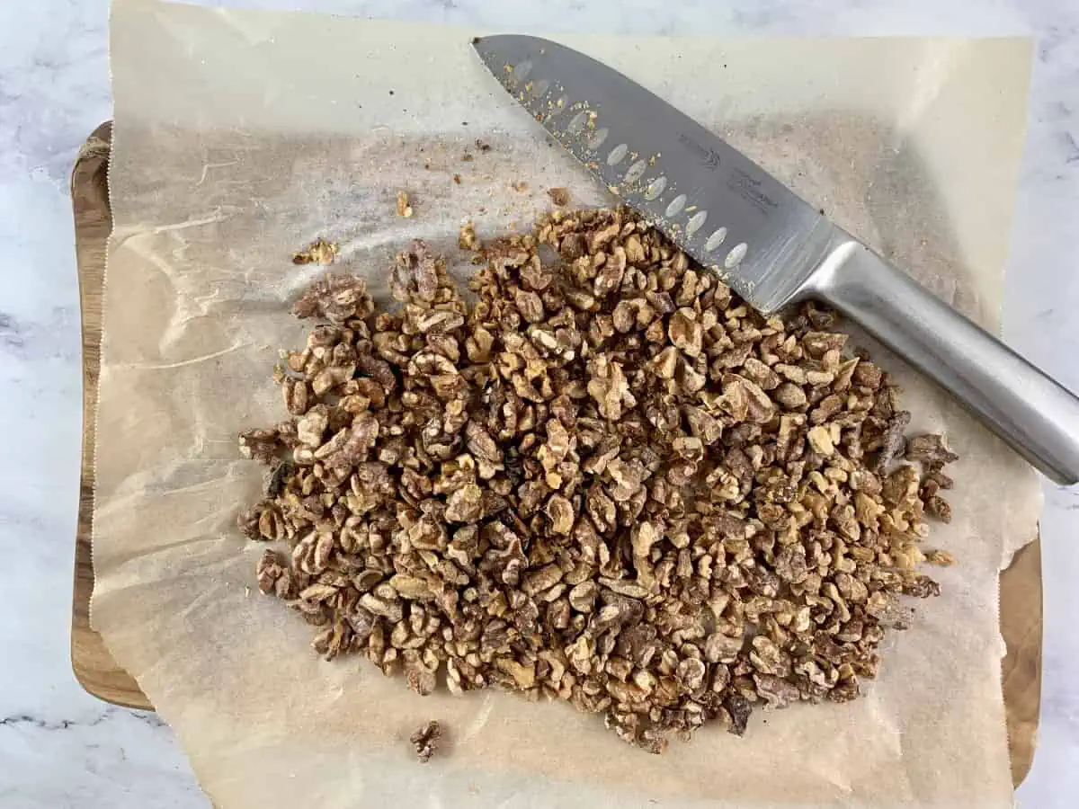 Chopping walnuts with a knife on a wooden board lined with baking paper.