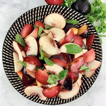White peach salad on a black patterned plate with plums and mint on the side.