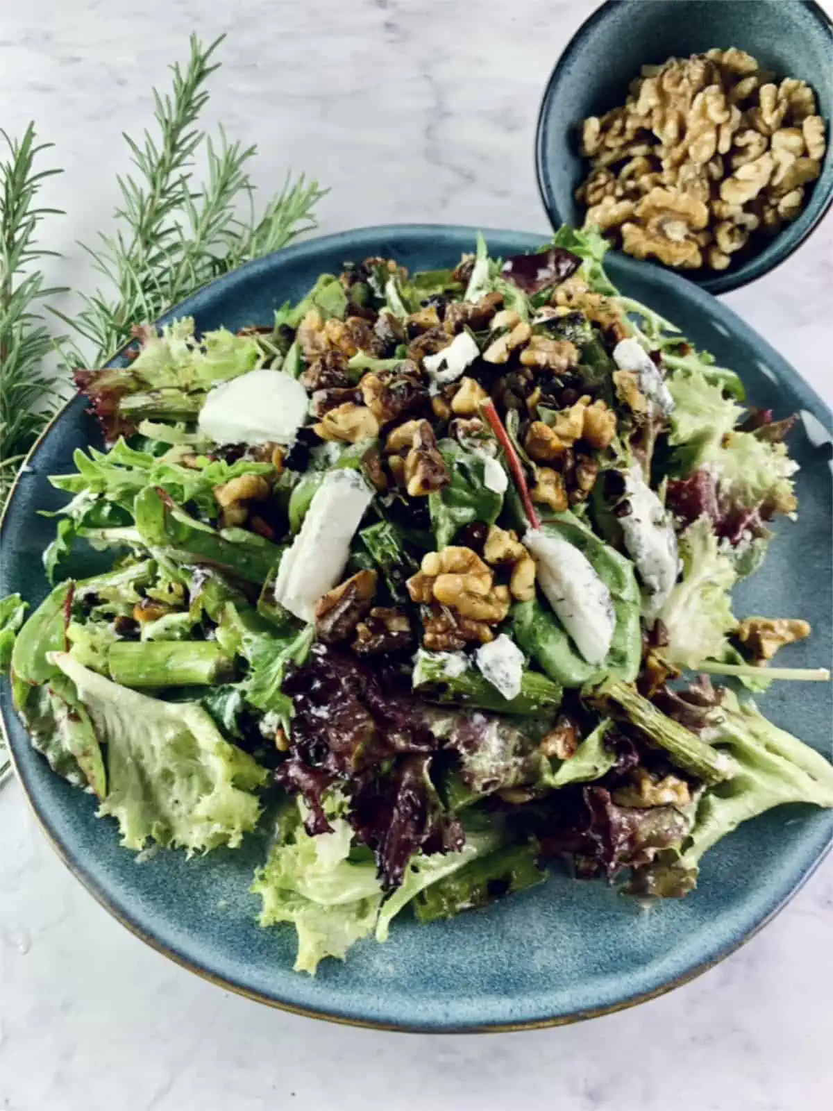 Barbecue asparagus salad on a blue plate with walnuts in top right corner and rosemary sprigs on the left.