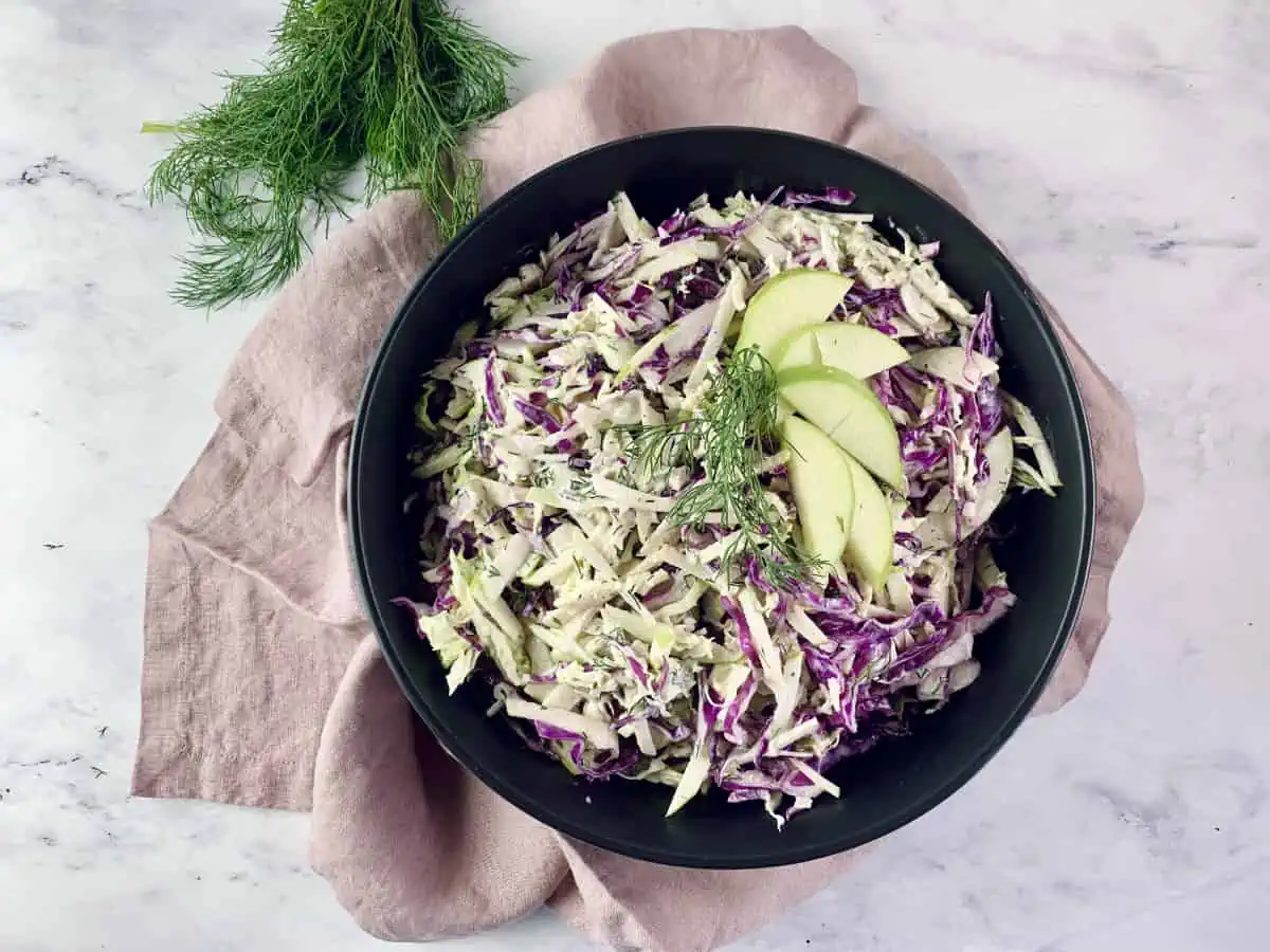 An aerial view of apple slaw in a black bowl with a pink linen napkin on the side. 