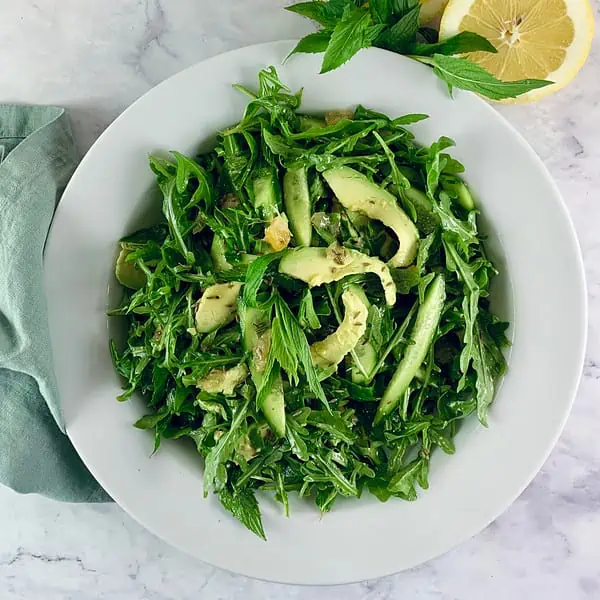Avocado rocket/arugula salad in a white bowl with lemon halves and mint sprigs on the side.