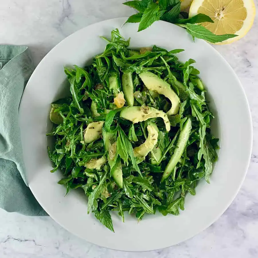 Avocado rocket/arugula salad in a white bowl with lemon halves and mint sprigs on the side.