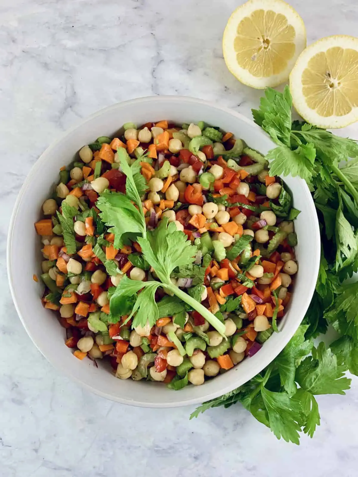 Vibrant garbanzo bean salad in a white bowl with celery leaves and lemon halves on the side.
