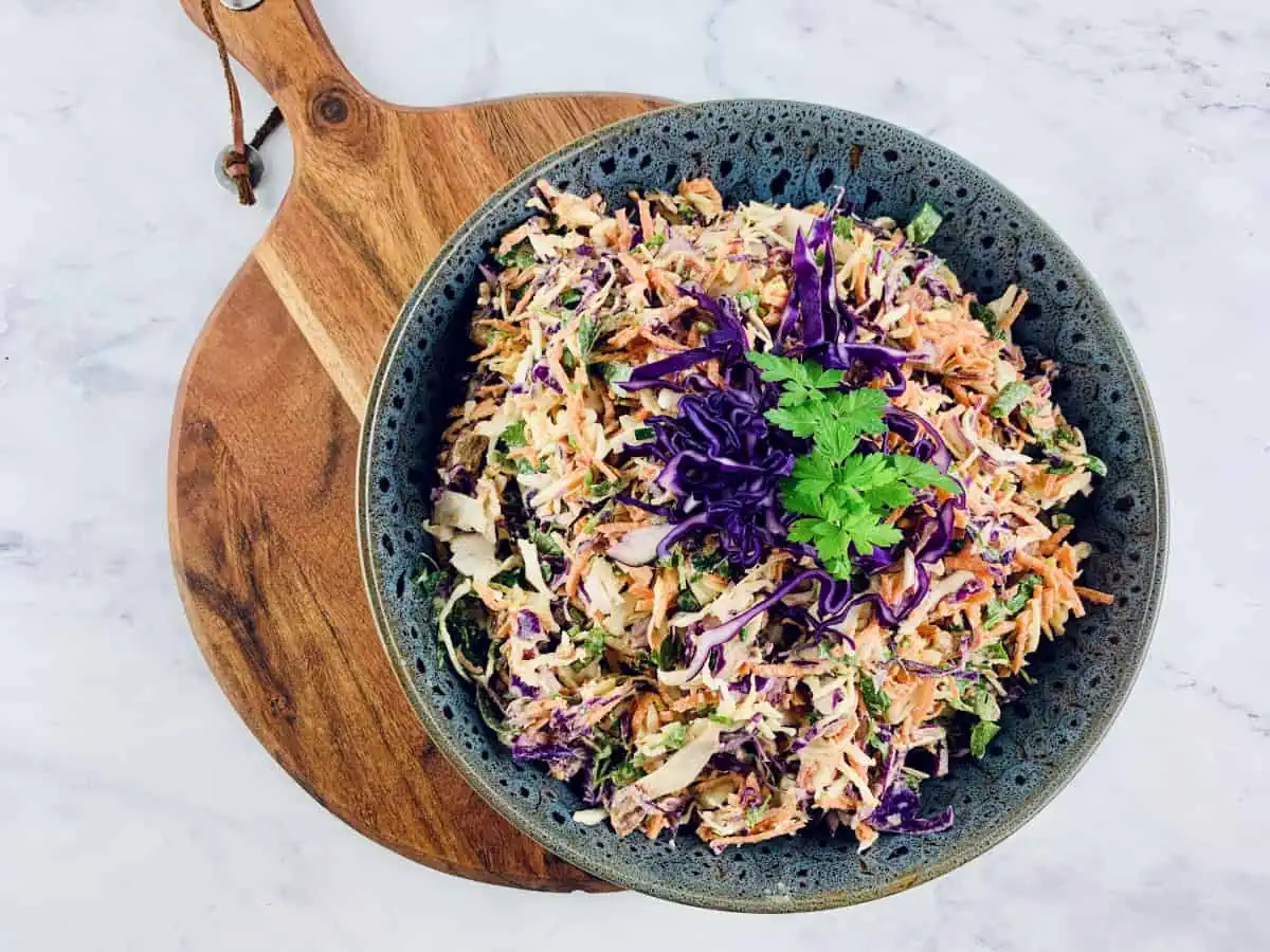 KETO COLESLAW IN A DARK GREY SALAD BOWL ON TOP OF A WOODEN BOARD WITH A RED CABBAGE PARSLEY GARNISH