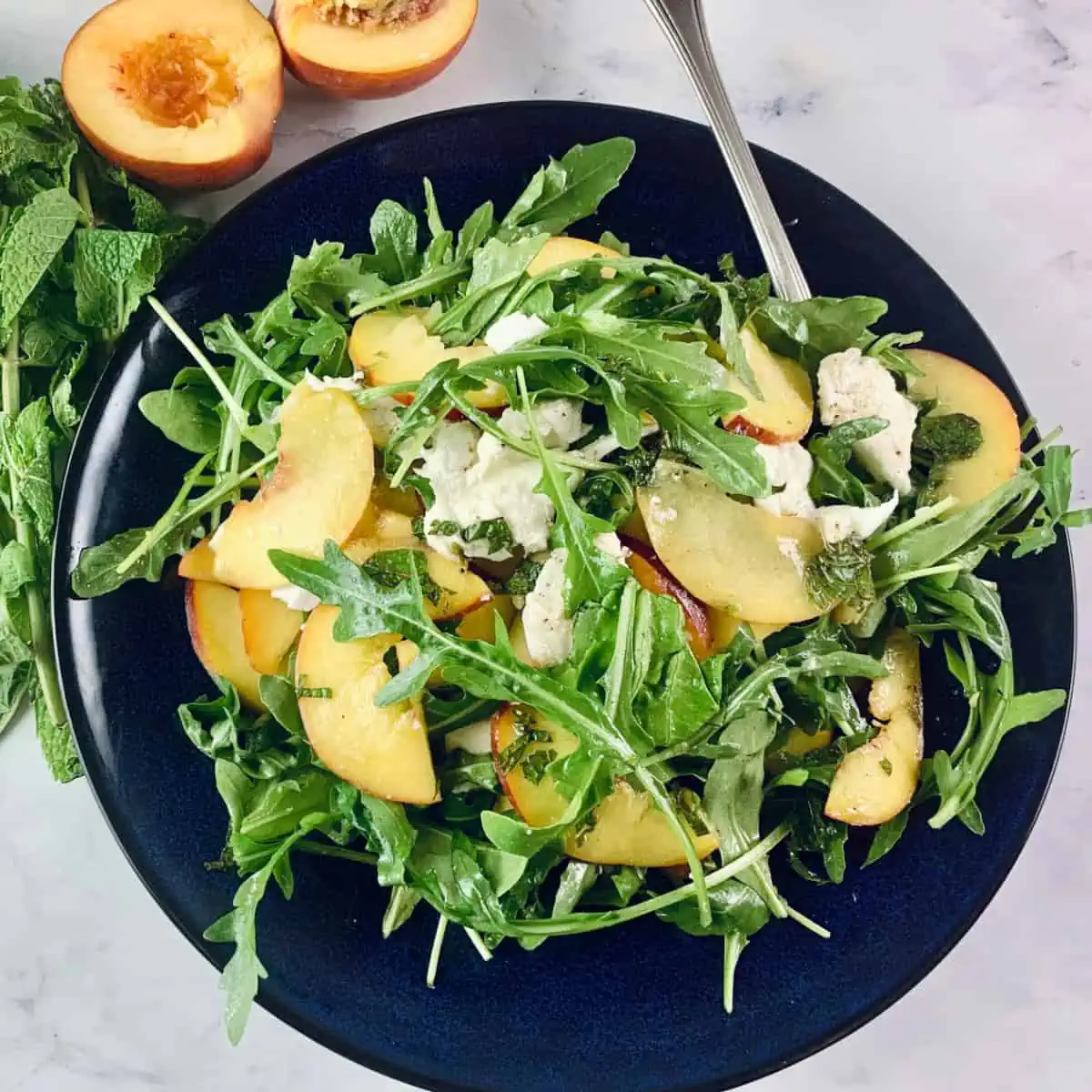 Peach salad on a navy plate with mint sprigs and halved peaches on the side.