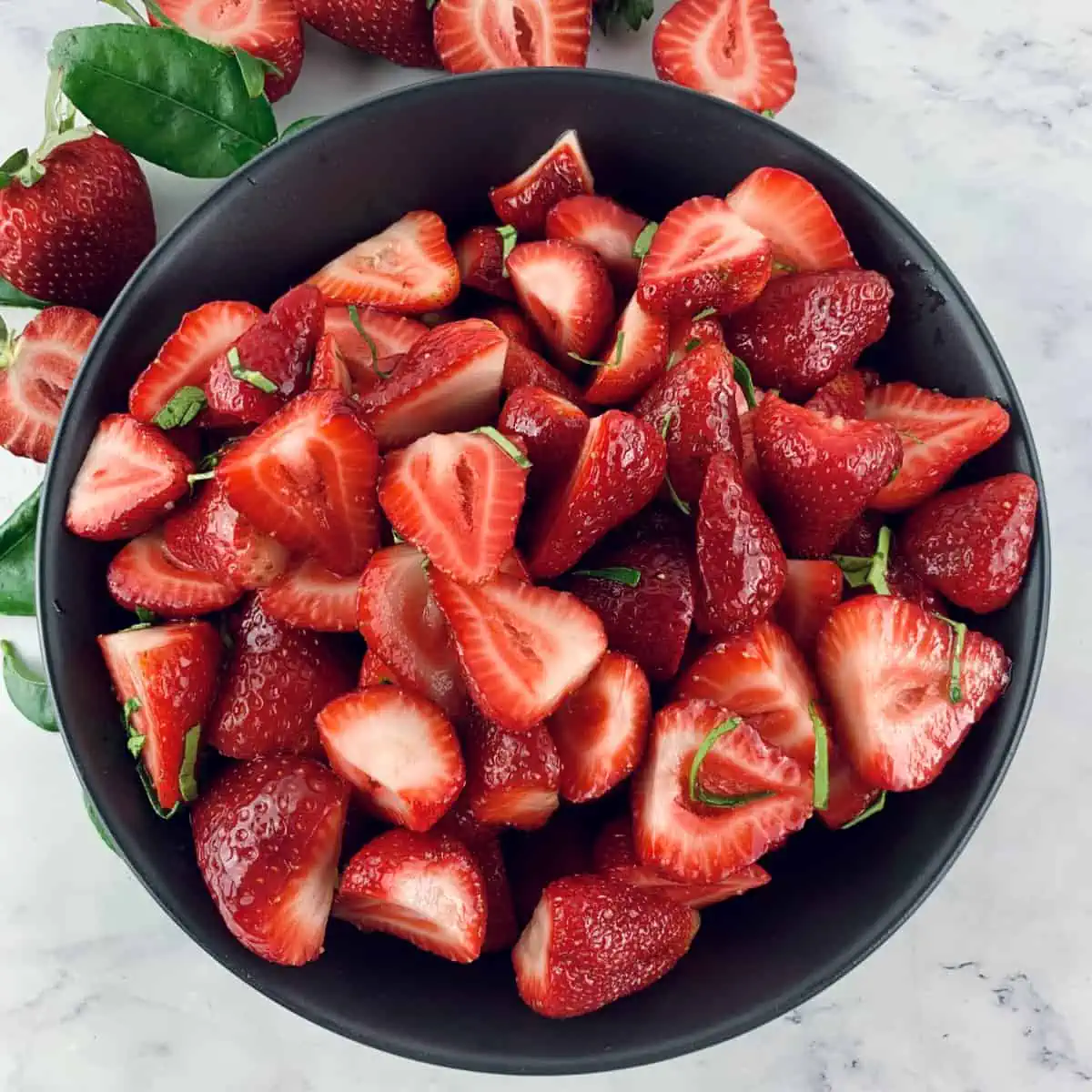 Strawberries and lime fruit salad in a black bowl with strawberries and lime leaves scattered around.