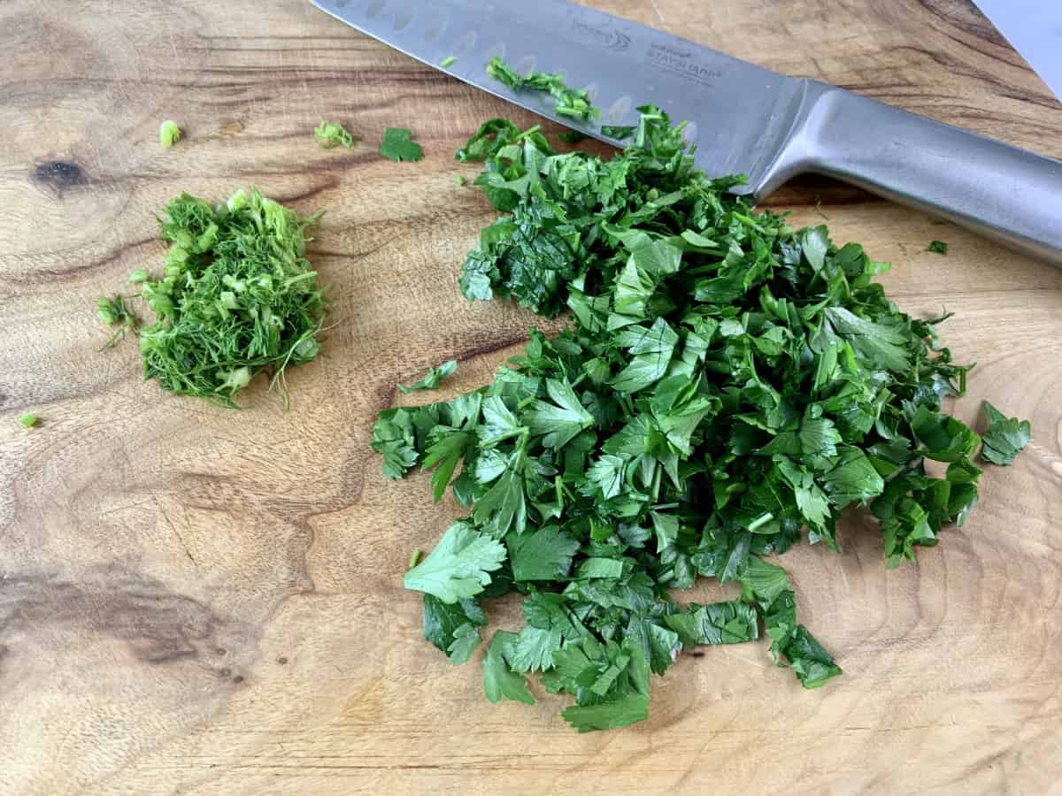 Chopping parsley and fennel fronds on a wooden board with a knife.