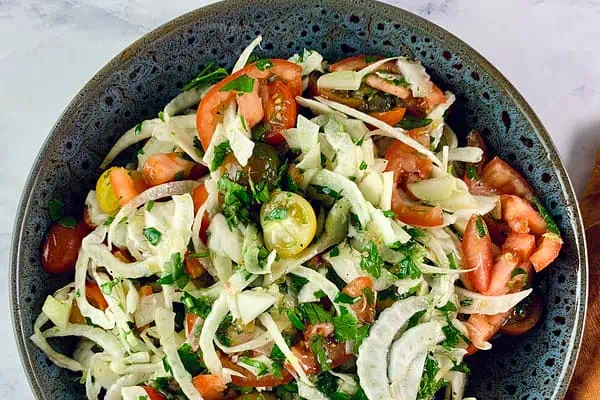 A close up of Fennel and Tomato Salad in a dark grey bowl with an orange linen napkin on the side.
