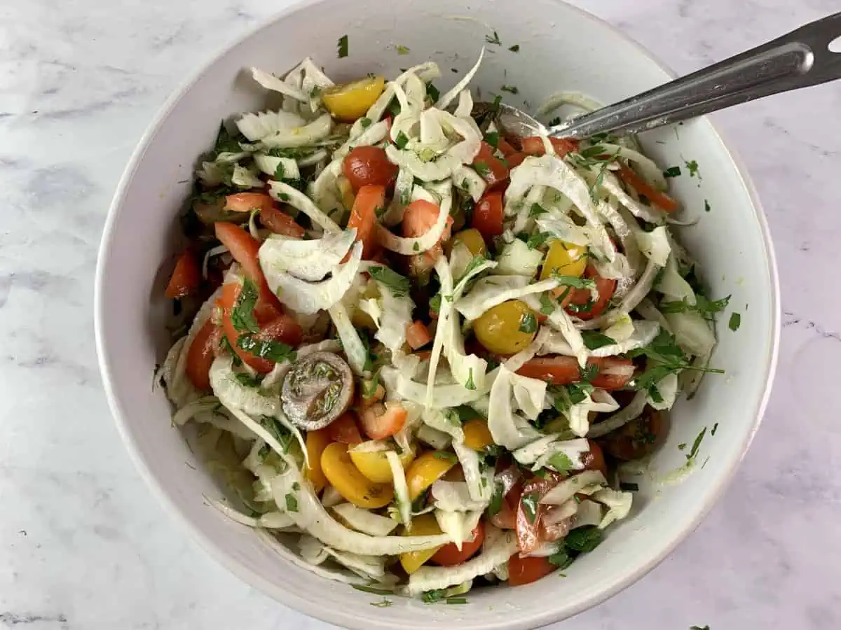 Mixing prepared fennel and tomato salad in a white bowl with a spoon.