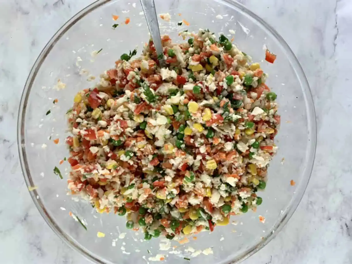 Mixing prepared riced cauliflower salad ingredients with mayo and lemon in a glass bowl.