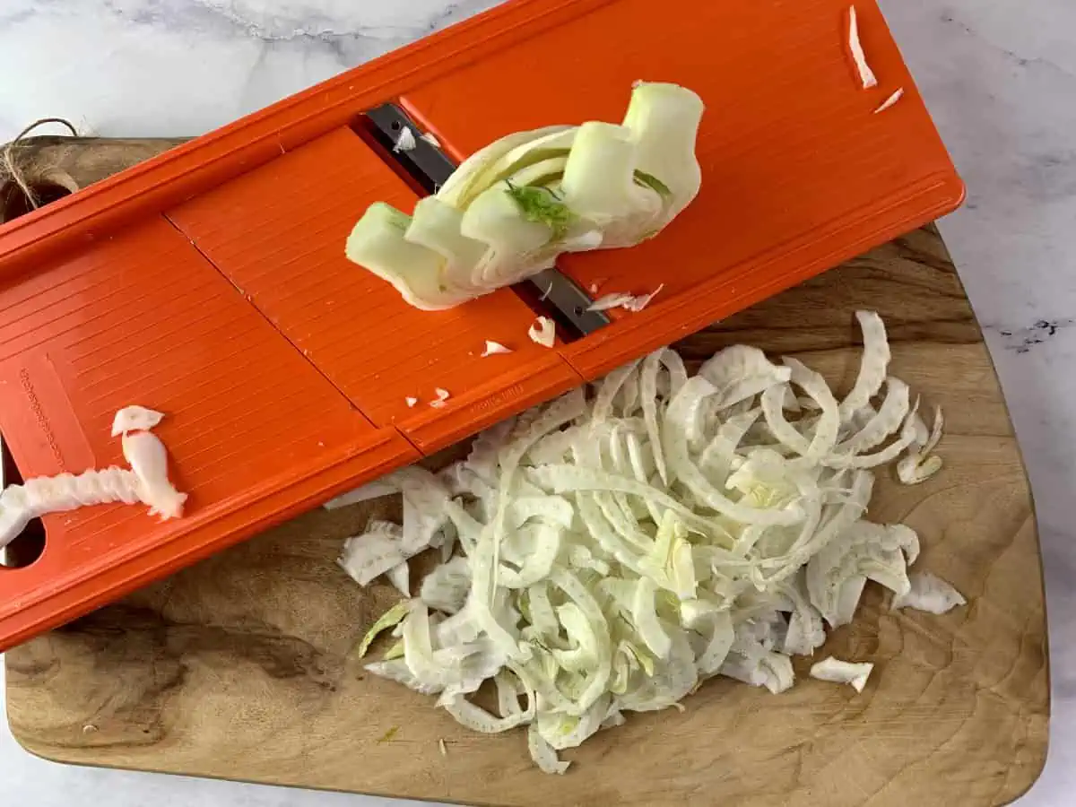 Shaving fennel with a vegetable slicer on a wooden board.