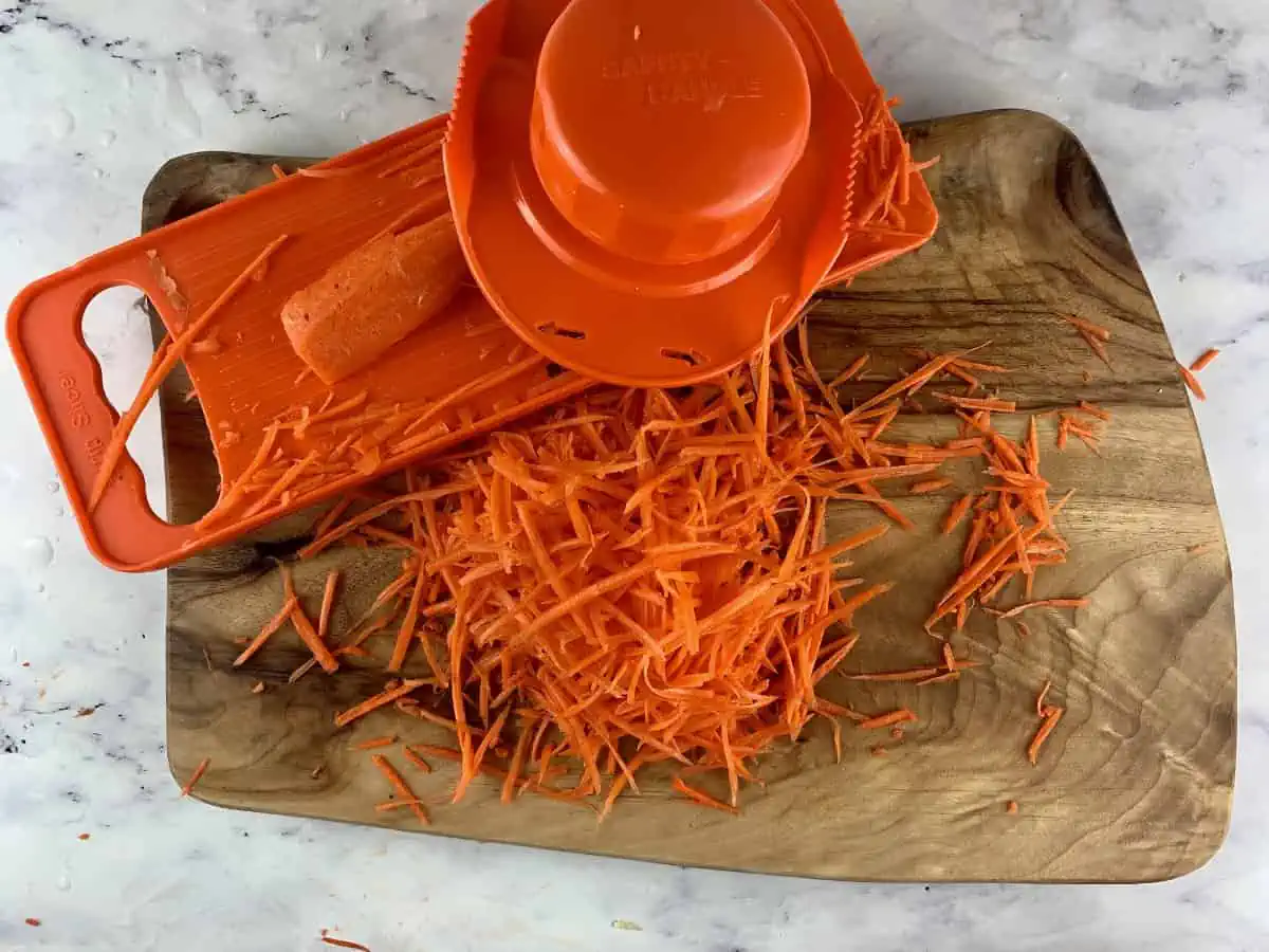 Carrots being julienned with an vegetable slicer on a wooden board.