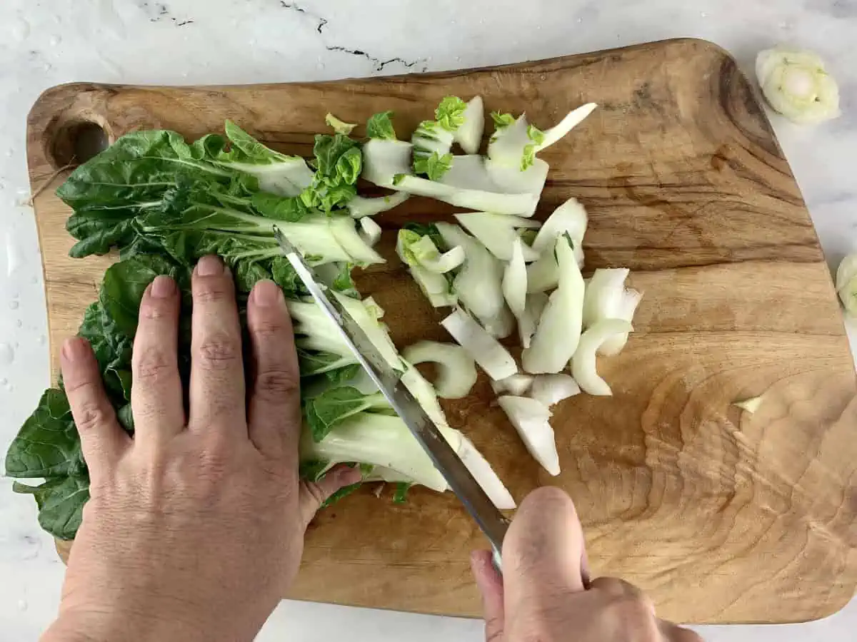 Hands diagonally slicing bok choy on a wooden board with a knife.