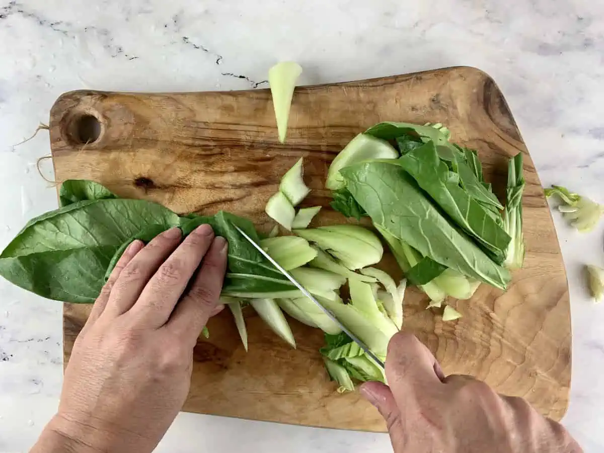 Hands diagonally slicing pak choy on a wooden board with a knife.